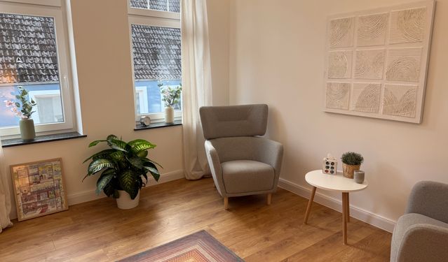 Heller, modern eingerichteter Therapieraum mit grünem Sofa, beigefarbenem Sessel, Holztisch und Pflanzen vor großen Fenstern.