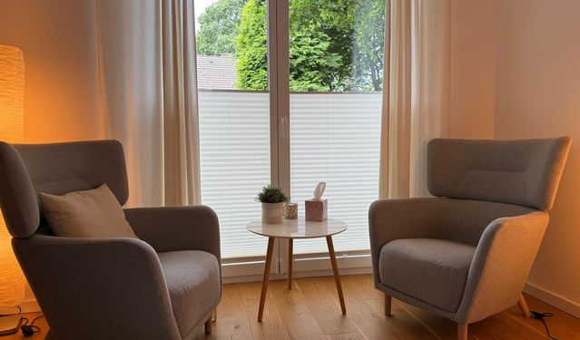 Heller, modern eingerichteter Therapieraum mit grünem Sofa, beigefarbenem Sessel, Holztisch und Pflanzen vor großen Fenstern.